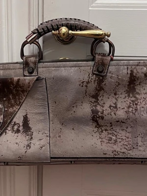 Gråbrun väska från Pucci - Unik väska i gråbrunt skinn med unika detaljer.Väskan har robusta handtag med snygga detaljer i mässingsfärg. Väskan har en avlång form och klassisk design, perfekt för dig som vill sticka ut. Märket heter Renzo Pucci och är handgjort i små upplagor i Italien. Den är numrerad se bilderna och därför väldigt intressant för den som samlar på väskor.
