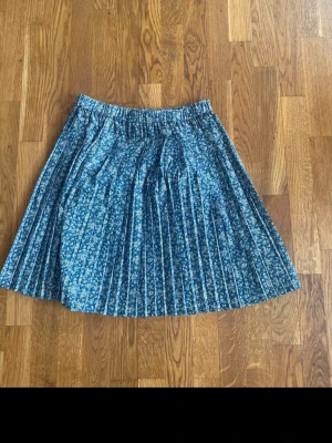 Blå plisserad blommig kortkjol - Säljer en blå kortkjol med vita små blommor och plisserad design. Kjolen har resår i midjan och är gjord i ett lätt och mjukt material, perfekt för vår och sommar. Superfin att matcha med en enkel topp eller stickad tröja.