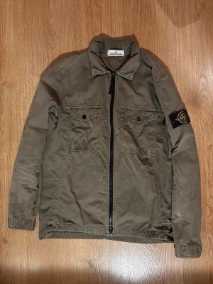 Stone Island Overshirt - Snygg olivgrön overshirt från Stone Island med dragkedja framtill, två bröstfickor med knappar och klassisk patch på vänster ärm. Tillverkad i slitstarkt bomullstyg och har en avslappnad passform med krage.
