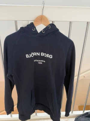 Marinblå hoodie från Björn Borg - Svart hoodie från Björn Borg med vit text på bröstet där det står 'BJÖRN BORG STOCKHOLM 1956'. Hoodien har huva med snörning och en stor magficka. Perfekt för en avslappnad och sportig stil. Storlek 170 i barn