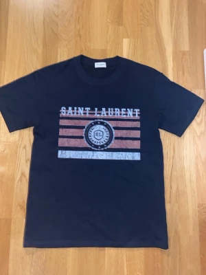 Svart Saint Laurent t-shirt - Snygg svart t-shirt från Saint Laurent med tryck framtill i vitt och rött. Klassisk rund halsringning och korta ärmar. Perfekt för dig som gillar streetstyle och vill ha något trendigt i garderoben.