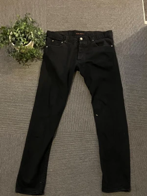 Svarta jeans från Nudie Jeans - Säljer ett par svarta nudie jeans. Jätte sköna och perfekt till dig som vill ha en avslappnad look, tyvärr lite trasiga som ner på sista bilderna 34/32 storlek 