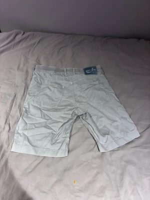 Ljusgråa shorts från Jacob cohen  - Säljer ett par ljusgråa shorts från Jacob Cohen  med klassisk femficksdesign och bälteshällor. De har knapp- och dragkedjestängning samt raka ben. Perfekta för varma dagar och enkla att matcha med olika stilar.