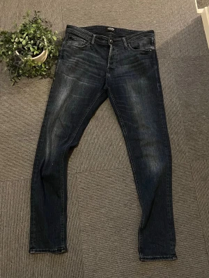 Mörkblå Jack and Jones jeans  - Snygga mörkblå jeans med slim fit passform och klassisk femficksdesign. Jeansen har subtila slitningar och kontrastsömmar för en modern look. Tillverkade i stretchigt denimtyg som ger skön komfort och bra passform.