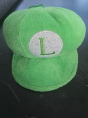 Grön Luigi-keps i plysch - Grön keps i mjukt plyschmaterial med en vit cirkel framtill och ett broderat grönt L. Klassisk form inspirerad av Luigi från tv-spel, perfekt som accessoar till cosplay eller maskerad.