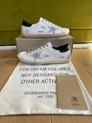 Golden Goose vita sneakers med stjärna - Säljer ett par vita Golden Goose sneakers med den ikoniska silverfärgade stjärnan på sidan och svarta detaljer vid hälen. Skorna har grå skosnören och en vintage-inspirerad, något smutsad look. Tillverkade i läder och levereras med dustbag och originalförpackning.