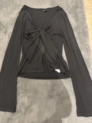 Svart långärmad topp med knyt - Snygg svart långärmad topp med stor knytning framtill som ger en trendig look. Toppen har en djup v-ringning och är tillverkad i ett mjukt, stretchigt material som sitter skönt på kroppen. Perfekt för dig som vill ha en stilren men ändå unik topp.