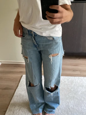 Ljusblå slitna baggy jeans - Säljer ett par ljusblå baggy jeans med flera stora slitningar och hål på både fram- och baksida av benen. Jeansen har en avslappnad passform och raka ben. Perfekta för en trendig och avslappnad stil.