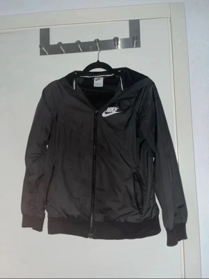 Svart Nike vindjacka med huva - Snygg svart vindjacka från Nike med vit logga på bröstet. Jackan har dragkedja framtill, två fickor med dragkedja och en praktisk huva. Resår vid ärmslut och nederkant ger en skön passform. Perfekt för blåsiga dagar och streetwear-stil.