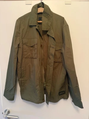 Olivgrön overshirt med fickor - Stilren olivgrön overshirt med två bröstfickor med lock och knapp, klassisk krage och knappstängning framtill. Tillverkad i ett lätt, slitstarkt material som passar perfekt för lager-på-lager. Patchdetalj nedtill på framsidan ger en cool touch.
