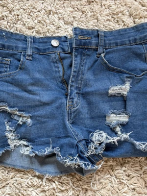 Två par slitna jeansshorts - Två par jeansshorts i olika blå nyanser, båda med hög midja och rejält slitna detaljer. Ena paret är mörkblått och det andra ljusblått, båda i klassisk denim med fransiga benslut och trasiga partier. Perfekta för en avslappnad sommarstil.