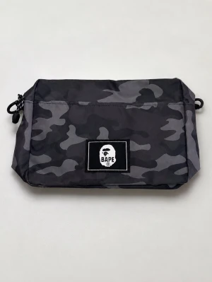 Svart kamouflageväska från BAPE - Snygg svart och grå kamouflageväska från BAPE med klassisk logga framtill. Väskan har dragkedja, meshficka på baksidan och praktiska snördetaljer på sidorna. Perfekt för att bära småprylar och accessoarer. Tillverkad i slitstarkt tyg. Aldrig använd innan sprillans ny.