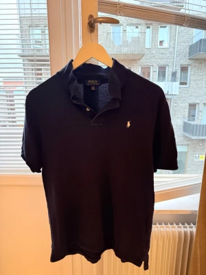 Mörkblå pikétröja från Polo Ralph Lauren - Klassisk Mörkblå pikétröja från Polo Ralph Lauren med den ikoniska broderade loggan på bröstet. Tröjan har korta ärmar, krage och knappar vid halsen. Tillverkad i mjuk bomull som känns skön mot huden.