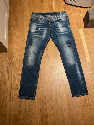 Blå slitna jeans från Dsquared2 - Säljer ett par blå jeans från Dsquared2 med slitningar, patchdetalj och ljusa tvättade partier. Jeansen har klassisk femficksmodell, knappgylf och raka ben. Materialet är denim i bomull. Perfekta för dig som gillar en trendig och avslappnad stil.