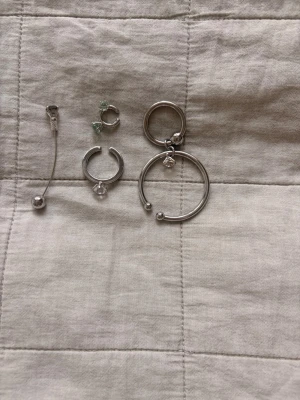 Mixade silvriga örhängen i olika former - Mixade silvriga smycken. 1 ring med vit diamant, 2 earcuffs en stor med vit diamant en liten med gröna diamanter, 1 örhänge med svart diamant.