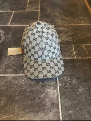 Gucci keps med GG-mönster - Snygg keps från Gucci i grått tyg med klassiskt GG-monogram över hela kepsen. På sidan finns en röd och blå rand som ger extra stilpoäng. Justerbar baktill för perfekt passform. Perfekt accessoar för dig som gillar exklusiv streetwear.