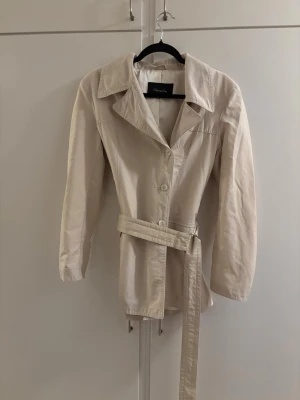 Beige trenchcoat med bälte - Stilren beige trenchcoat med klassisk krage och knäppning framtill. Jackan har ett matchande bälte i midjan för snygg passform och långa ärmar. Perfekt för vår och höst, tillverkad i ett lätt tyg som ger en clean look.