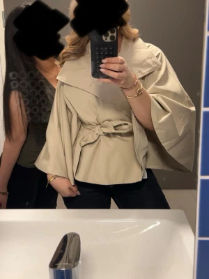 Beige trenchcoat med knytskärp - Stilren beige trenchcoat med vida ärmar och knytskärp i midjan. Jackan har en oversized krage och ett mjukt, följsamt tyg som ger en avslappnad men chic look. Perfekt för dig som vill ha en trendig och unik trenchcoat till din garderob.