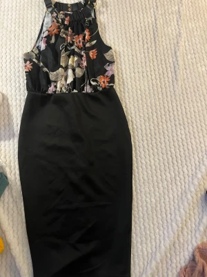 Svart blommig klänning AX Paris - Snygg svart klänning från AX Paris med blommigt mönster upptill och enkel svart kjoldel. Klänningen har halterneck med guldfärgad ringdetalj och är ärmlös. Perfekt för dig som vill ha en stilren men ändå unik look. Prislappen kvar 