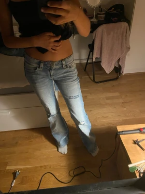 Ljusblå bootcut jeans med slitningar - Säljer ett par ljusblå bootcut jeans med slitningar på knäna och snygga detaljer på bakfickorna. Jeansen har låg midja och klassisk femficksdesign. Perfekta för en avslappnad och trendig look.