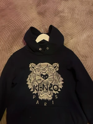 Svart Kenzo hoodie med tigertryck - Svart hoodie från Kenzo med ikoniskt tigerbroderi och texten 'KENZO PARIS' på bröstet. Klassisk huva med dragsko och känguruficka framtill. Perfekt för dig som gillar streetwear och vill sticka ut med en snygg detalj.