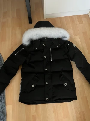 Svart dunjacka med pälskantad huva - Snygg svart dunjacka med stor huva som har vit pälskant. Jackan har flera fickor med dragkedjor och knappar, samt detaljer i silverfärg. Puffig modell med dragkedjor vid ärmslut och nederkant. Perfekt för kalla vinterdagar.