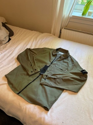 Olivgrön overshirt från Stone Island - Snygg olivgrön overshirt från Stone Island med dragkedja framtill, bröstficka med lock och klassisk patch på ärmen. Overshirten har en avslappnad passform och är tillverkad i ett lätt, slitstarkt material som passar perfekt till lager-på-lager-stil.