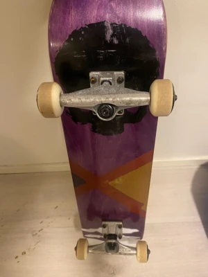 Skateboard från NKD Sports - Säljer en skateboard med lila och svart grafisk design på undersidan och svart grepptejp med NKD Sports-logga på ovansidan. Brädan har vita hjul och silverfärgade truckar. Perfekt för dig som vill cruisa eller öva tricks.