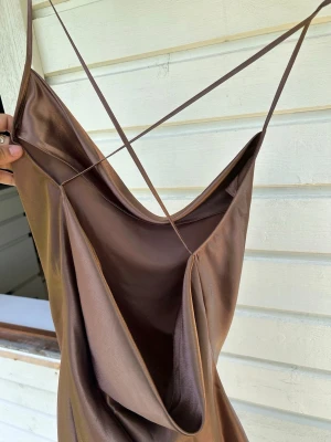 Brun långklänning i satin - Snygg brun långklänning med tunna axelband och draperad urringning. Klänningen har en slät, glansig yta och är tillverkad i satinliknande material. Perfekt för dig som gillar minimalistisk och elegant stil.