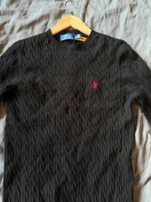 Svart kabelstickad tröja Polo Ralph Lauren - Kabelstickad svart tröja från Polo Ralph Lauren med rund halsringning och den klassiska röda logotypen broderad på bröstet. Tröjan har långa ärmar och ribbade muddar vid ärmslut och nederkant. Perfekt för dig som gillar stilrena och tidlösa plagg. Kan gå ner lite i pris vid en snabb affär. Den är m men sitter som en xs/s
