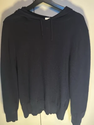 Svart cashmere hoodie med huva - En enkel mörkblå cashmere hoodie med huva och dragsnören. Modellen har långa ärmar och ribbade muddar vid ärmslut och nederkant. Skick är 10/10 och inga defekter på tröjan, passar perfekt till alla tillfällen. Priset kan diskuteras vid snabbaffär! 