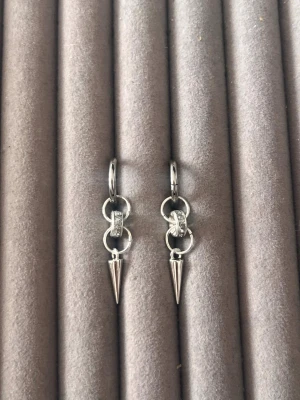 Silvriga örhängen med konformad hängare - Säljer ett par coola silvriga örhängen med små ringar och en konformad hängare. Örhängena har en minimalistisk design med en liten detalj av gnistrande stenar i mitten. Perfekta för dig som gillar edgy accessoarer.
