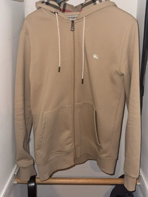 Beige hoodie från Burberry med dragkedja - Snygg beige hoodie från Burberry med dragkedja, klassiskt rutigt foder i huvan och vita dragsnören. Har två stora fickor framtill och liten Burberry-logga på bröstet. Perfekt för en stilren och avslappnad look.