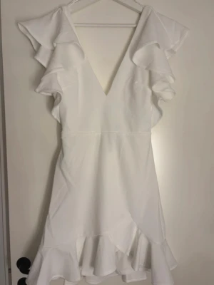 Vit volangklänning med öppen rygg - Superfin vit klänning med volangdetaljer vid axlar och fåll, djup v-ringning och öppen rygg med snygg knäppning. Klänningen är kort och har en feminin, flowy passform. Perfekt för sommarens alla tillfällen!