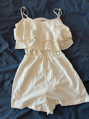 Vit somrig jumpsuit med volanger - Söt vit jumpsuit med tunna axelband och dubbla volanglager och broderade blommönster. Resår i midjan för skön passform och korta ben. Perfekt för varma dagar! Kontakta mig om ni har några frågor💕