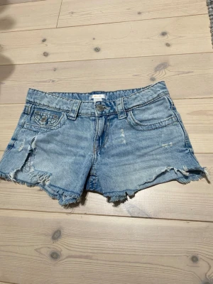 Blå jeansshorts med broderade fickor - Säljer ett par ljusblå jeansshorts med fransig kant och slitningar. Shortsen har broderade bakfickor med snygga detaljer och knappstängning. Klassisk femficksmodell och bälteshällor. Perfekta för sommaren!