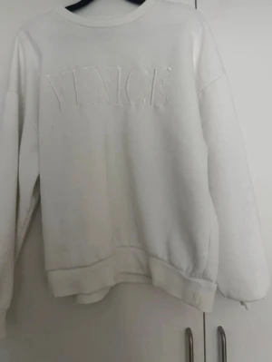 Vit sweatshirt med VENICE-tryck - Säljer en oversized vit sweatshirt med upphöjt VENICE-tryck på bröstet. Tröjan har rund halsringning, långa ärmar och ribbade muddar vid ärmslut och nederkant. Perfekt för en chill och avslappnad stil. Materialet känns mjukt och skönt mot huden.