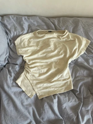Beige omlottopp med kort ärm - Säljer en beige omlottopp med korta ärmar och avslappnad passform. Toppen har en snygg omlottdetalj i sidan som ger en cool twist till din outfit. Perfekt att matcha med jeans eller kjol för en trendig look.