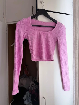 Ljusrosa croppad topp med mönster - Trendig ljusrosa croppad topp med lång ärm och geometriskt mönster i tyget. Toppen har en bred, rund ringning och är figurnära i modellen. Perfekt att styla med höga jeans eller kjol.