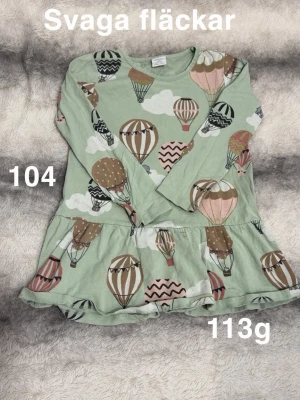 Grön tunika med luftballonger - Söt grön tunika med volangkant och lång ärm, täckt av ett lekfullt mönster med luftballonger i brunt, rosa, beige och vitt. Perfekt för dig som gillar färg och roliga prints.🌸 Svaga fläckar. Storlek 104