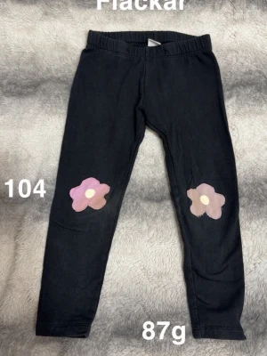 Svarta leggings med blommor - Svarta leggings med resår i midjan och två söta blommor i rosa och lila på knäna. Perfekta för dig som gillar en lekfull och färgglad stil. 🌸 Fläckar. Storlek 104