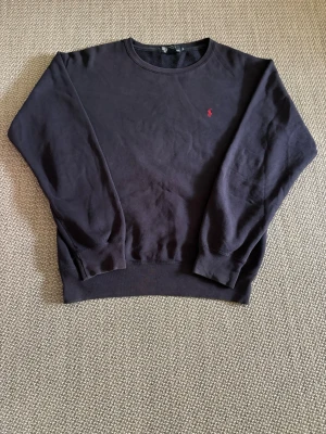 Mörkblå stickad tröja Polo Ralph Lauren - Mörkblå stickad tröja från Polo Ralph Lauren med rund halsringning och diskret röd logga broderad på bröstet. Tillverkad i 82% bomull och 18% polyester. Klassisk passform och ribbade muddar vid ärmslut och nederkant.