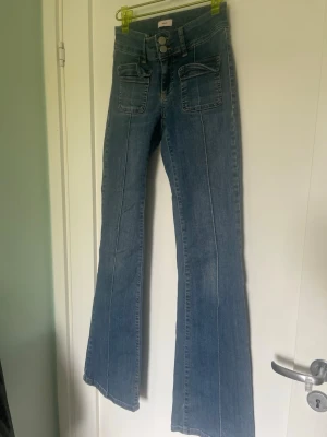 Blå utsvängda jeans med fickor - Säljer ett par blå jeans med utsvängda ben och markerad pressveck framtill. Jeansen har dubbla knappar i midjan och stora fyrkantiga fickor fram. Materialet är klassisk denim och modellen ger en retrovibe.