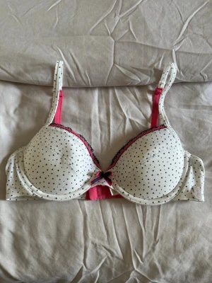 Gullig Vit bh med svarta prickar och rosett - Super Söt bh i vitt med små svarta prickar (polkadot mönster), vadderade kupor och bygel!😍Axelbanden är justerbara i en vacker rosa färg och kuporna har spetskant i rosa och svart framtill. Mellan kuporna sitter en dekorativ rosett i rosa och svart🩷Perfekt för dig som gillar unika, söta, och bekväma bh:ar. Materialet är mjukt och skönt, passar super för alla plagg! Använd några gånger, inga fel. En bra bh för dig men små bröst eller en nybörjar bh✨Hör av vid funderingar eller bud!❤️