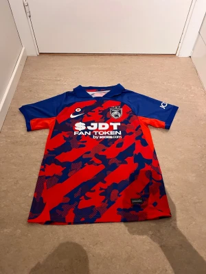 Röd och blå fotbollströja JDT - Snygg fotbollströja i rött och blått med kamouflagemönster och korta ärmar. Tröjan har JDT-logga på bröstet och tryck med 'FAN TOKEN by socios.com' framtill. Tillverkad i lätt och ventilerande material, perfekt för träning eller match.