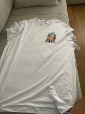 Vit Casa Blanca t-shirt med tryck - Säljer en vit t-shirt med färgglatt tryck från Casa Blanca. Motivet visar en solnedgång, palmer och vaser i grönt, rosa och gult. T-shirten har rund hals och korta ärmar. Perfekt för dig som gillar grafiska prints och vill sticka ut.