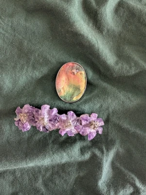 Lila blomformad hårklämma & brosch - Säljer en lila hårklämma med blommor i plast och glittriga detaljer samt en oval brosch med skimrande pastellfärger i rosa, grönt och gult. Perfekt för att ge din frisyr en unik och färgglad touch.