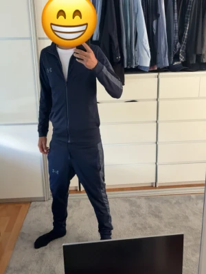 Mörkblått Underarmour tracksuit - Mörkblått underarmour tracksuit i storlek S. Både byxor och en zip tröja. Perfekt för träning och daglig användning. Var ett litet hål på höger knä som är hyfsat lagat. Man ser knappt det när man använder det och tänker inte på det.