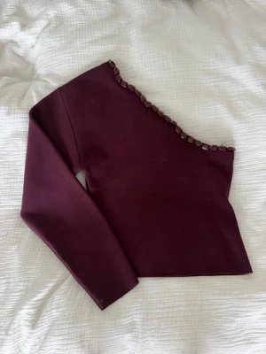 Vinröd oneshoulder topp med detaljer - Snygg vinröd oneshoulder topp med långa ärmar och dekorativa stenar längs halsringningen. Toppen har en croppad passform och är perfekt för dig som vill sticka ut. Materialet känns mjukt och stretchigt, troligen polyester eller syntet. Använd en gång