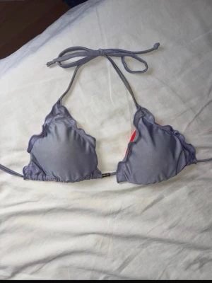 Vändbar bikiniöverdel i rött och lila - Säljer en vändbar trekantsbikini med volangkant. Ena sidan är lila och den andra sidan är röd. Bikinin har knyt i nacken och ryggen, vilket gör den enkel att justera. Perfekt för sommardagar på stranden eller vid poolen. Den är lite smutsig vid banden och guldknappen i mitten har rostat lite. 